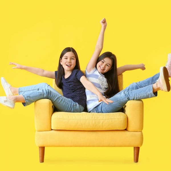 Two girls sitting on a yellow chair with arms in the air.